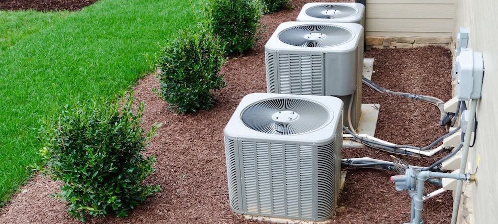 Three split-type outdoor units or condensers positioned side by side outside, adjacent to a well-manicured lawn.